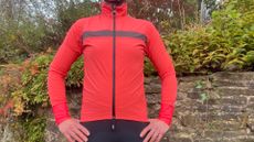 The Santini Guard Neo Rain Jacket in bright pink on a woman's torso standing in front of a wall and greenery 